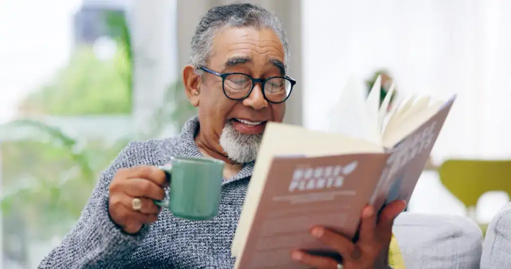 senior man reading book and holding mug after joining a book club for seniors at Clarendale Arcadia in Phoenix, AZ